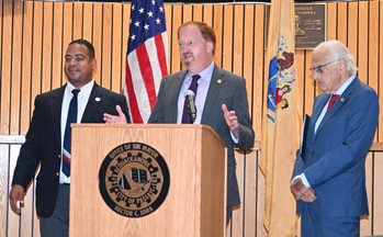 NJTPA Chair John Bartlett speaks at a podium, standing beside Congressman Bill Pascrell and Passaic Mayor Hector Lora.